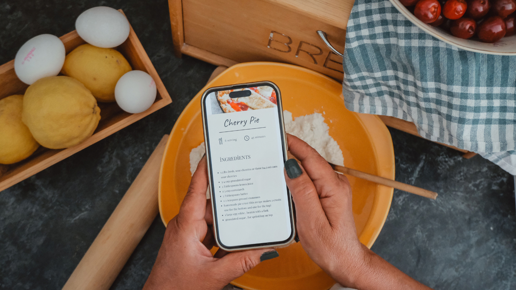 Smartphone antigo em suporte na bancada da cozinha exibindo aplicativo de receitas ao lado de ingredientes.